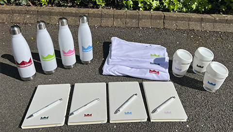 Company Rebranding With Multiple Colour Logos On Bottles, Notebooks, Pens, Coffee Cups, And T-Shirts